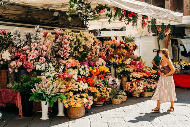Een bloemenkraam op een markt, vol emmers en manden met bloemen, en een vrouw die rondkijkt